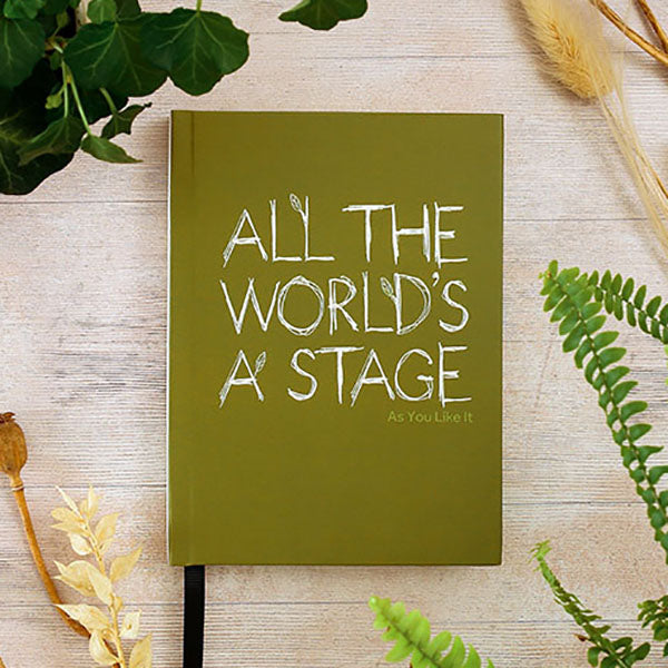 Green notebook with 'All the World's a Stage' quote on a wooden surface with plants.