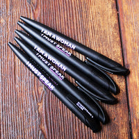 Black pens with 'I AM A WOMAN I MUST SPEAK' text on a wooden surface