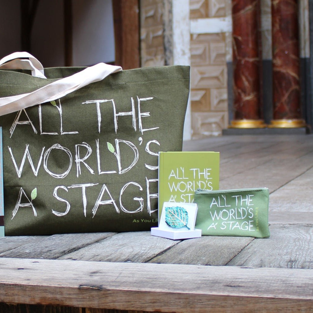 Wood panel stage floor with As You Like It sage green colour merchandise, left from right, baseball cap, paperback manga, tote bag, leaf brooch, hardback journal, purse