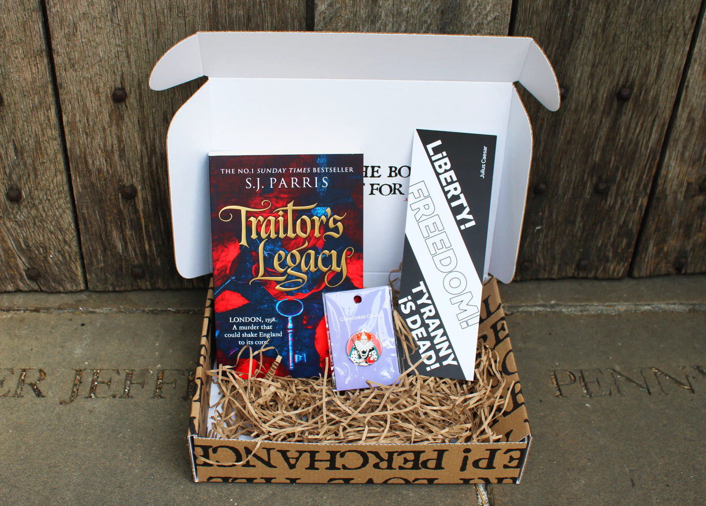 An open white subscription book box filled with shredded paper contains the historical novel Traitor's Legacy by S.J. Parris, standing upright beside a black-and-white bookmark featuring the quote "Liberty! Freedom! Tyranny is Dead!" from Julius Caesar by William Shakespeare. In front is a small round enamel pin depicting Queen Elizabeth I. The items are neatly arranged against a rustic wooden background.