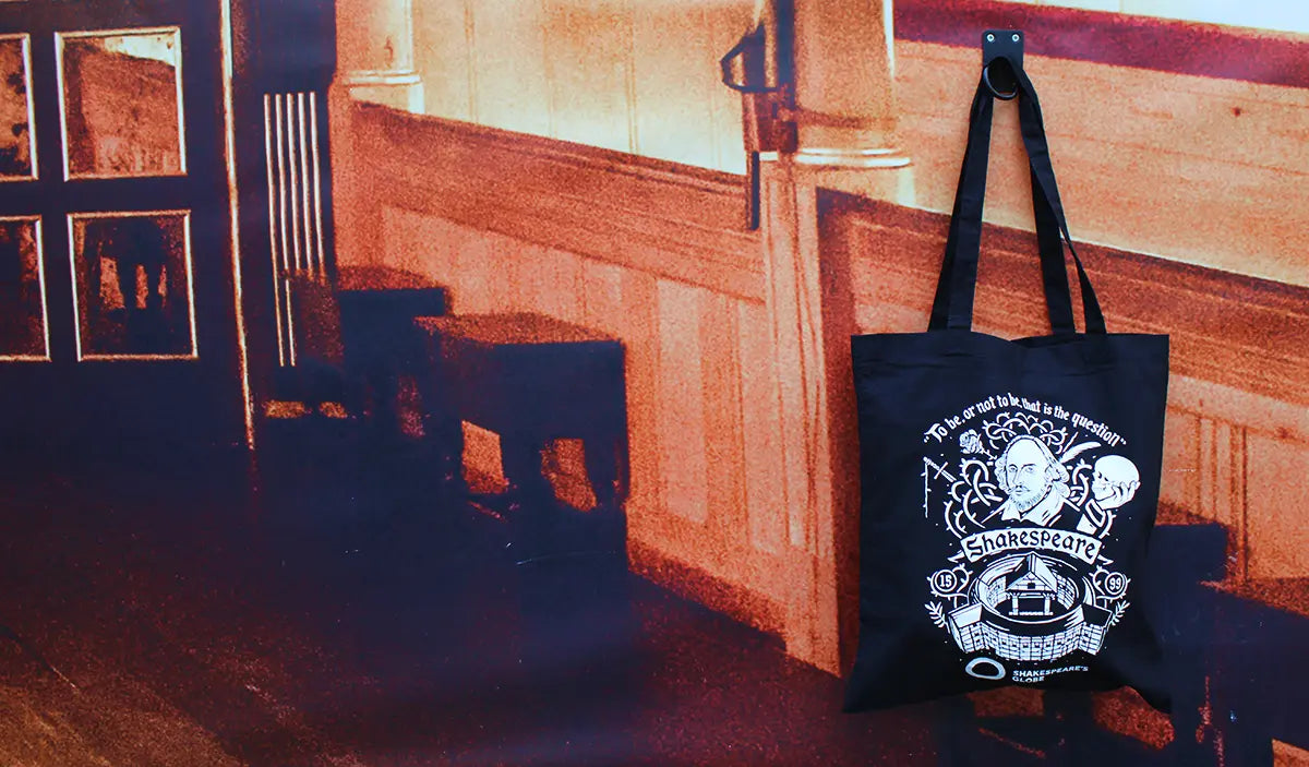 Black Shakespeare's Globe tote bag featuring Shakespeare, the Globe Theatre and the quote "To be, or not to be, that is the question", hanging in front of a printed backdrop depicting a room inside Shakespeare's Globe Theatre.