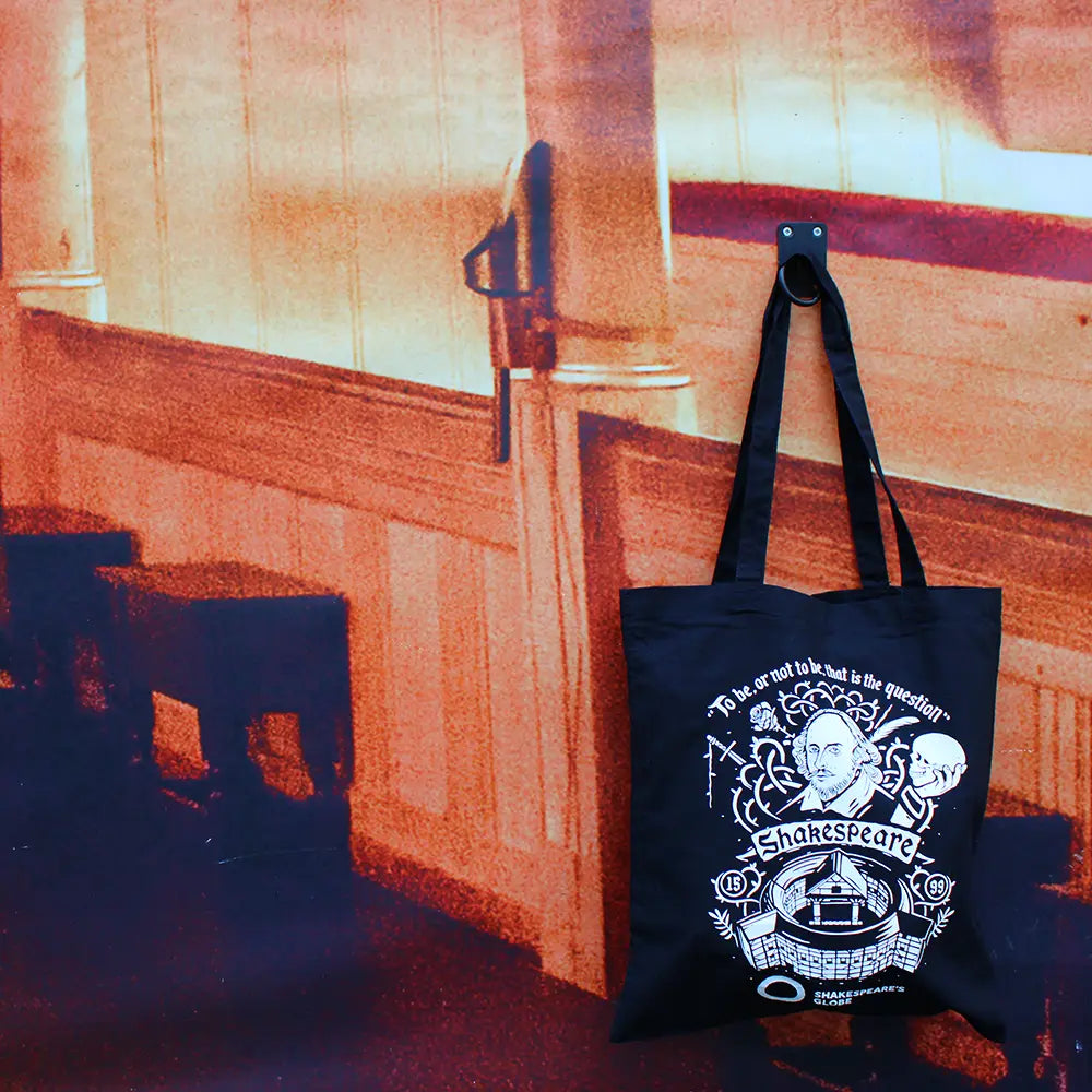 Black Shakespeare's Globe tote bag featuring Shakespeare, the Globe Theatre and the quote "To be, or not to be, that is the question", hanging in front of a printed backdrop depicting a room inside Shakespeare's Globe Theatre.