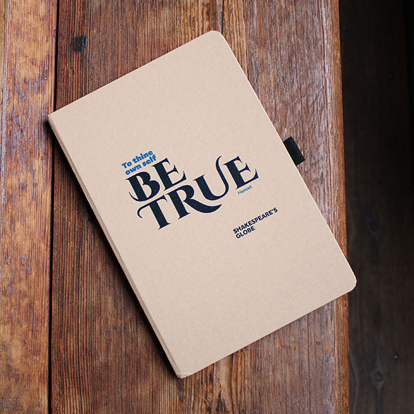 Notebook with 'Be True' quote by Shakespeare's Globe on a wooden background