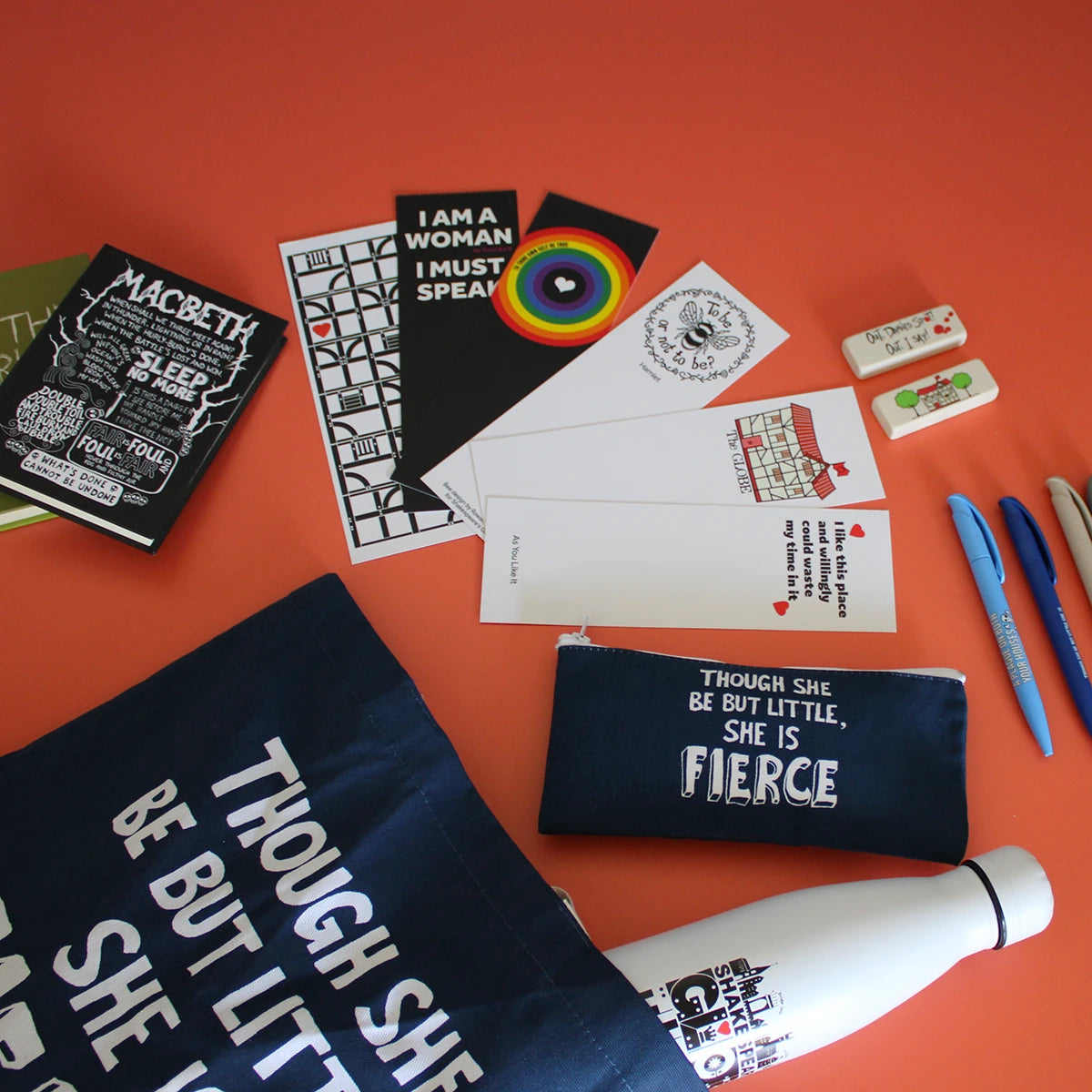 hakespeare-inspired back-to-school stationery and accessories on a bright orange background, featuring a navy pencil case and tote bag printed with the A Midsummer Night’s Dream quote “Though she be but little, she is fierce,” a Macbeth notebook, literary postcards and bookmarks, colourful pens, erasers and a reusable water bottle.