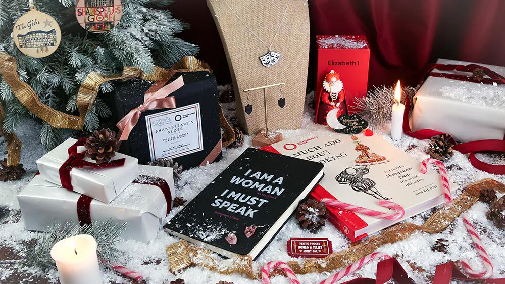 Festive Shakespeare-themed Christmas display featuring an ‘I Am a Woman, I Must Speak’ notebook, a theatre mask necklace, Romeo and Juliet brooch, Shakespeare jewellery, an Elizabeth I decoration, and Much Ado About Cooking, surrounded by wrapped gifts, candles, candy canes, pinecones, ribbons, and artificial snow.