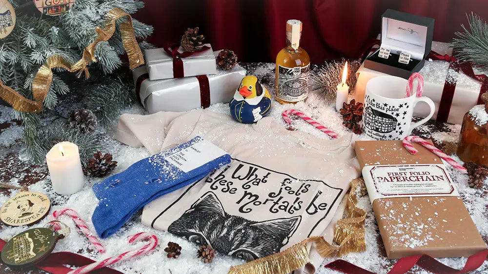 Festive Shakespeare-themed Christmas display featuring a cat T-shirt, blue socks, a Shakespeare rubber duck, Globe Theatre mug, Shakespeare cufflinks, a bottle of traditional mead, and a First Folio paperchain kit, surrounded by wrapped gifts, candles, candy canes, pinecones, ribbons, and artificial snow