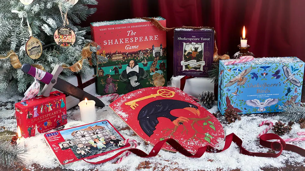 Festive Shakespeare-themed Christmas display featuring The Shakespeare Game, Shakespeare Tarot, Shakespeare’s Birds puzzle, Shakespeare’s Dramatic Gods game, a Globe Theatre kit, a dagger, and a red shield, surrounded by candles, ribbons, pinecones, and artificial snow.