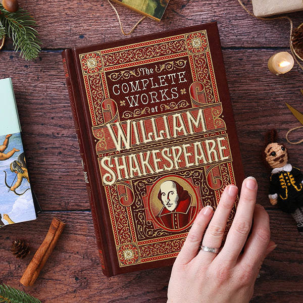 Decorative book titled 'The Complete Works of William Shakespeare' held by a hand on a wooden surface.