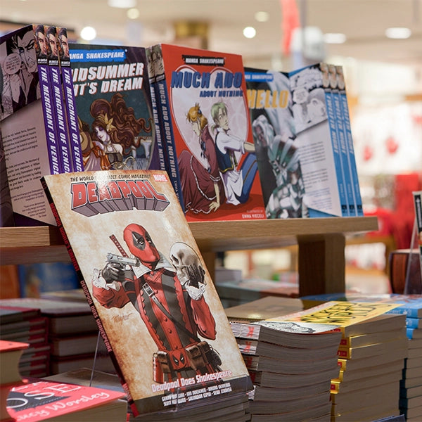 Image of 2 tiers of paperback graphic novels on a wooden table with Deadpool Does Shakespeare in the foreground