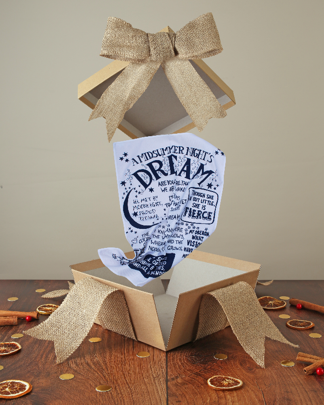 White cotton tea towel with royal blue print, popping out of kraft box with bow on wood base with Christmas decor