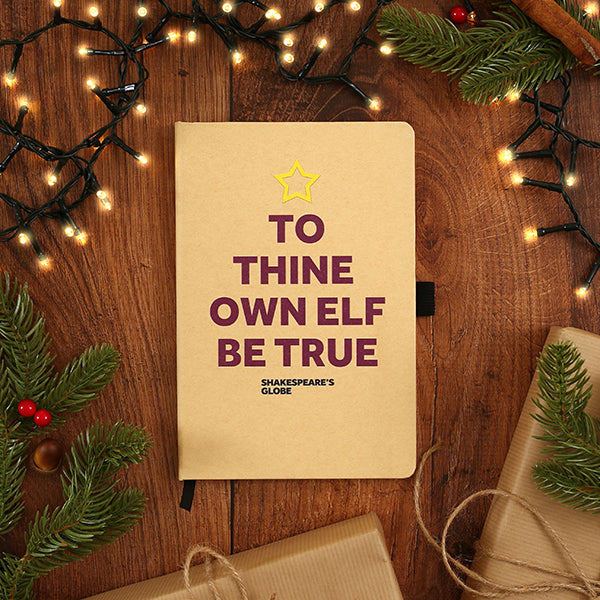 Notebook with 'To Thine Own Elf Be True' text on a wooden surface with Christmas decorations.