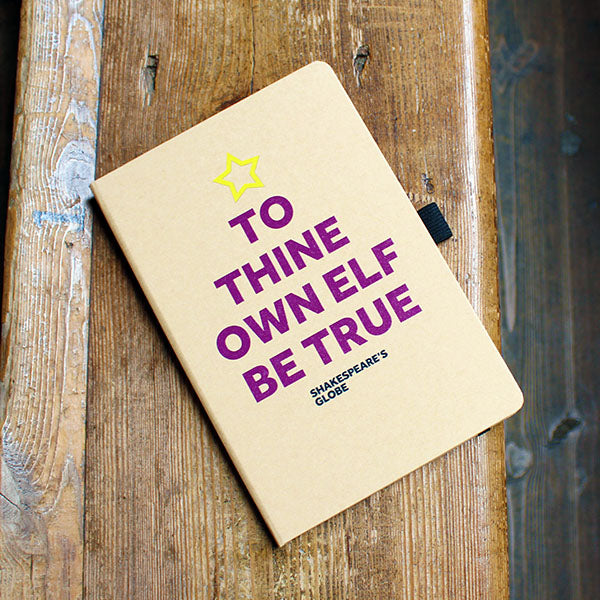 Kraft notebook with plum coloured block text in the shape of a Christmas tree with yellow star on top, lying on a wooden bench.