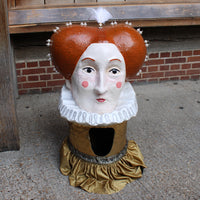 A painted headdress of Elizabeth I with a white ruffled collar, orange-brown hair, pink cheeks, and a gold ruffled base, photographed outdoors against a brick and wood background.