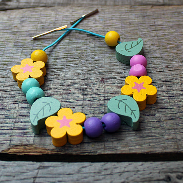 Wooden bracelet kit with green leaves, yellow flowers, and yellow, purple, pink, and teal balls on teal stretch string