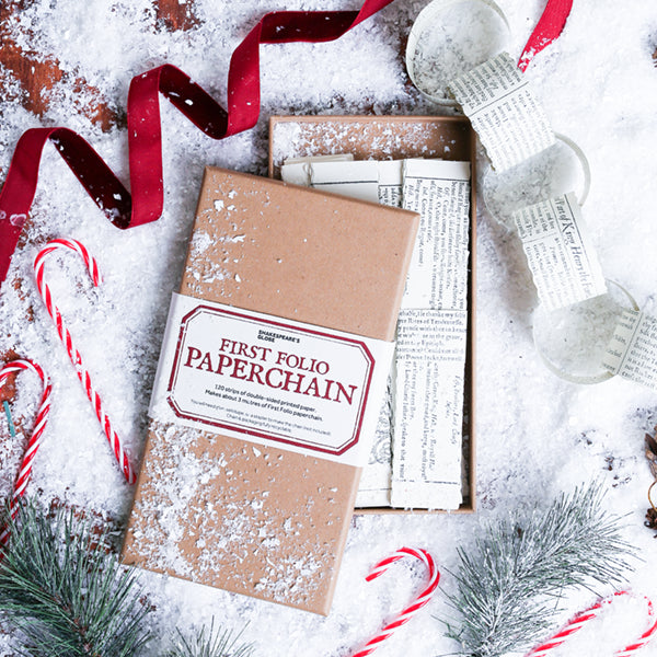 A flat-lay of a 'First Folio Paperchain' gift set, featuring a kraft paper box with a paper label, and a partially unfolded paper chain, all styled with red ribbon, candy canes, and pine branches on a snowy white background.