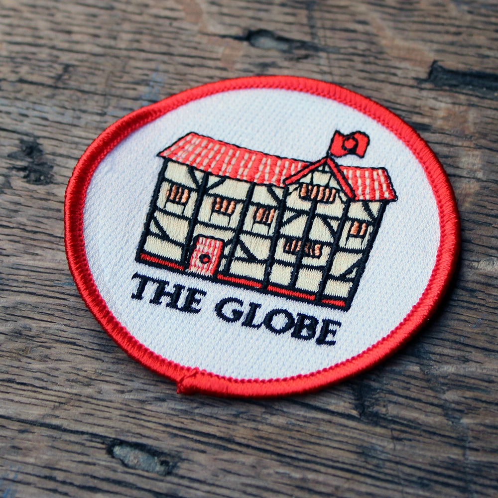 
                      
                        White circular embroidered badge with red border, featuring black, beige and red cartoon Globe in the centre
                      
                    