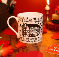 A white porcelain mug with a black and white illustration of the Globe Theatre and the quote 'I like this place' printed on it on a wooden surface with a red background
