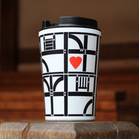 A white reusable travel cup with a black lid sits on a wooden surface. The tumbler features a black representation of the Globe Theatre's wooden frame with a small red heart in the centre, set against a blurred warm brown background.
