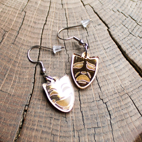Pair of gold coloured mask-shaped earrings on a wooden surface