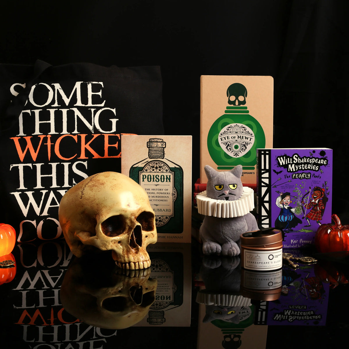 Halloween display featuring a ‘Something Wicked This Way Comes’ tote bag, a skull, glowing pumpkins, a black cat figurine, a lit candle, spooky bottles, and a William Shakespeare Mysteries book against a dark background