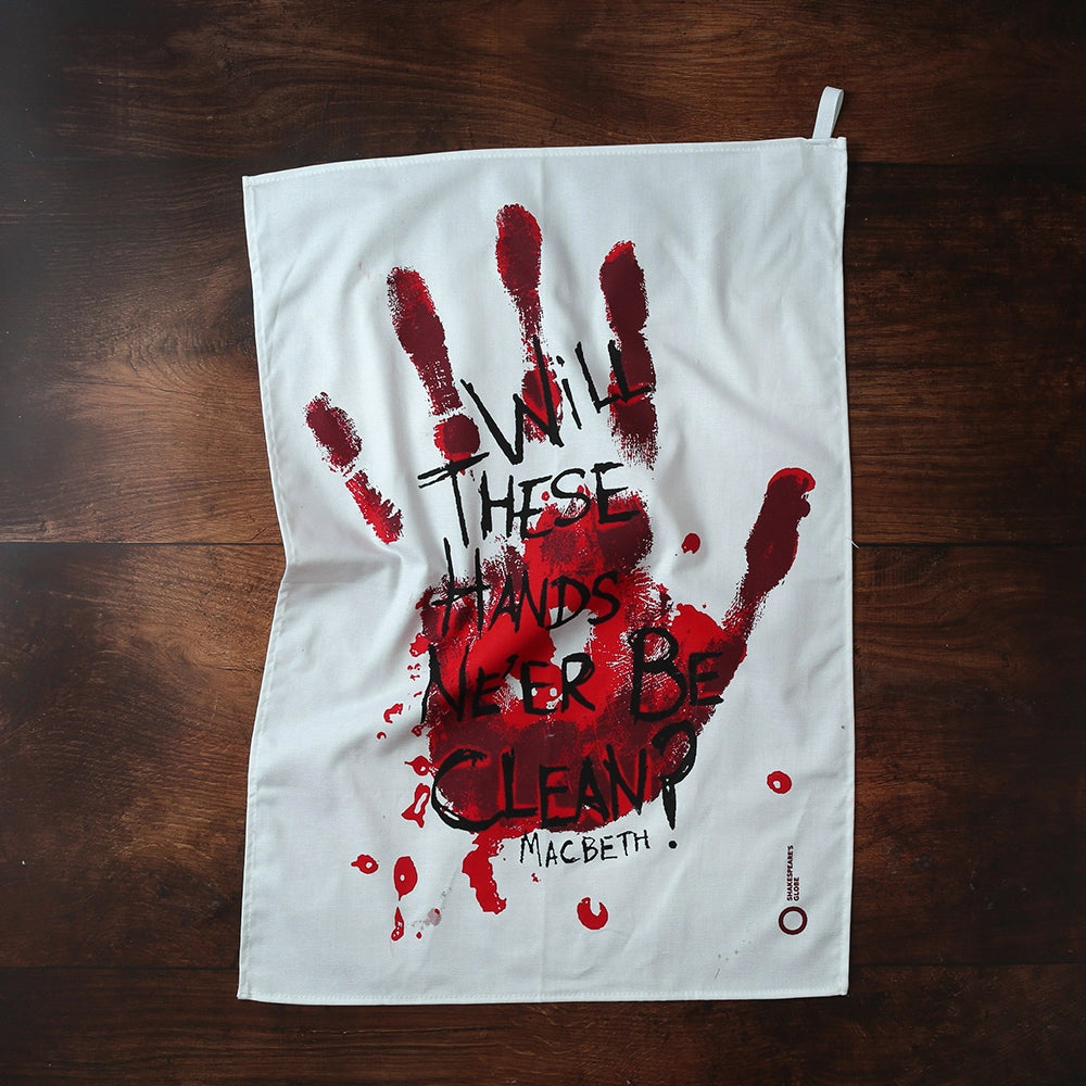 White cotton tea towel printed with a large bloody hand-print in reds. Over the hand-print, printed in black is a quote from Shakespeare play, Macbeth (Will these hands ne'er be clean?) The lettering is in an angry, hand-drawn style.