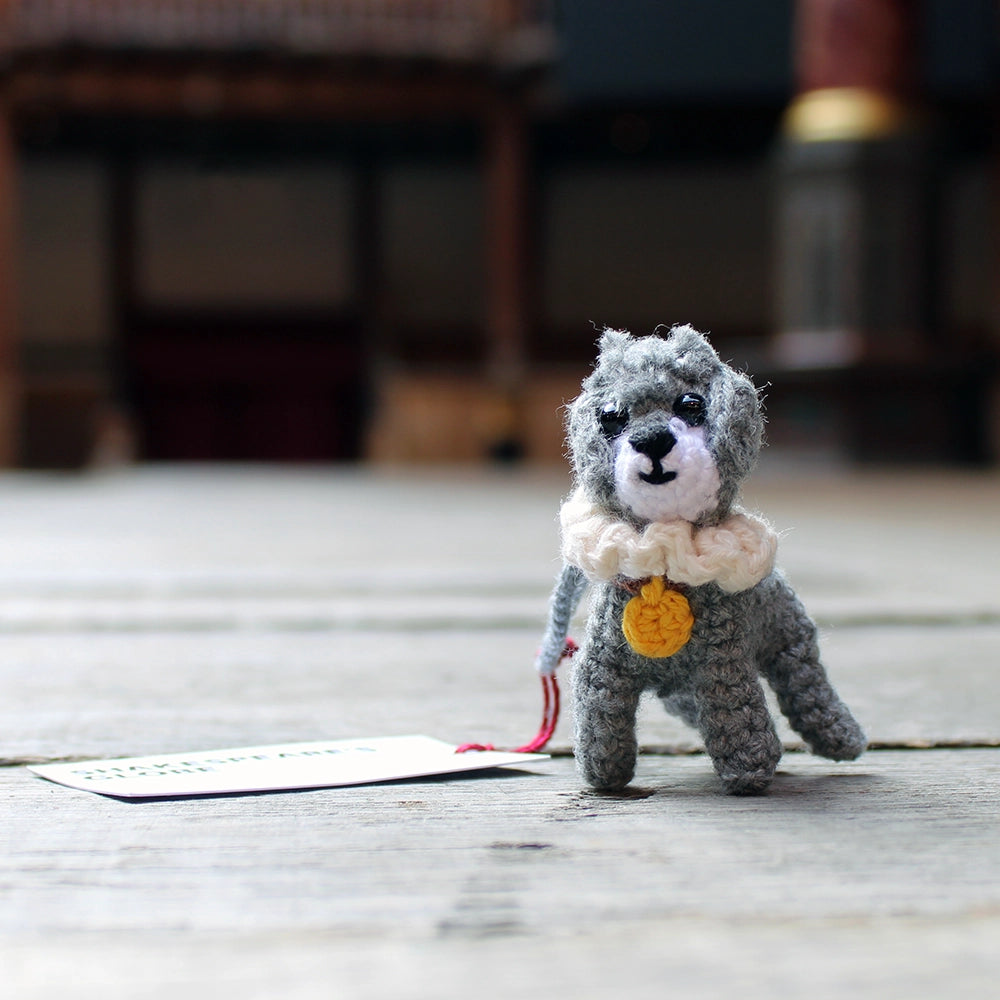 
                      
                        crocheted dog made from grey yarn with a white muzzle and black eyes. The dog wears a white ruff collar with a yellow tag
                      
                    