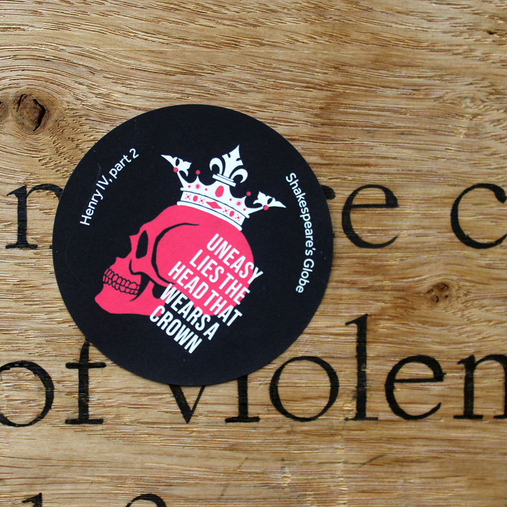 Black circular sticker with red skull wearing white crown, with white text overlayed, stuck on wood