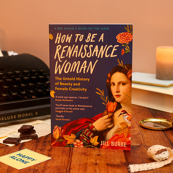 Book titled 'How to Be a Renaissance Woman' by Jill Burke on a wooden surface with a vintage typewriter and candles in the background.
