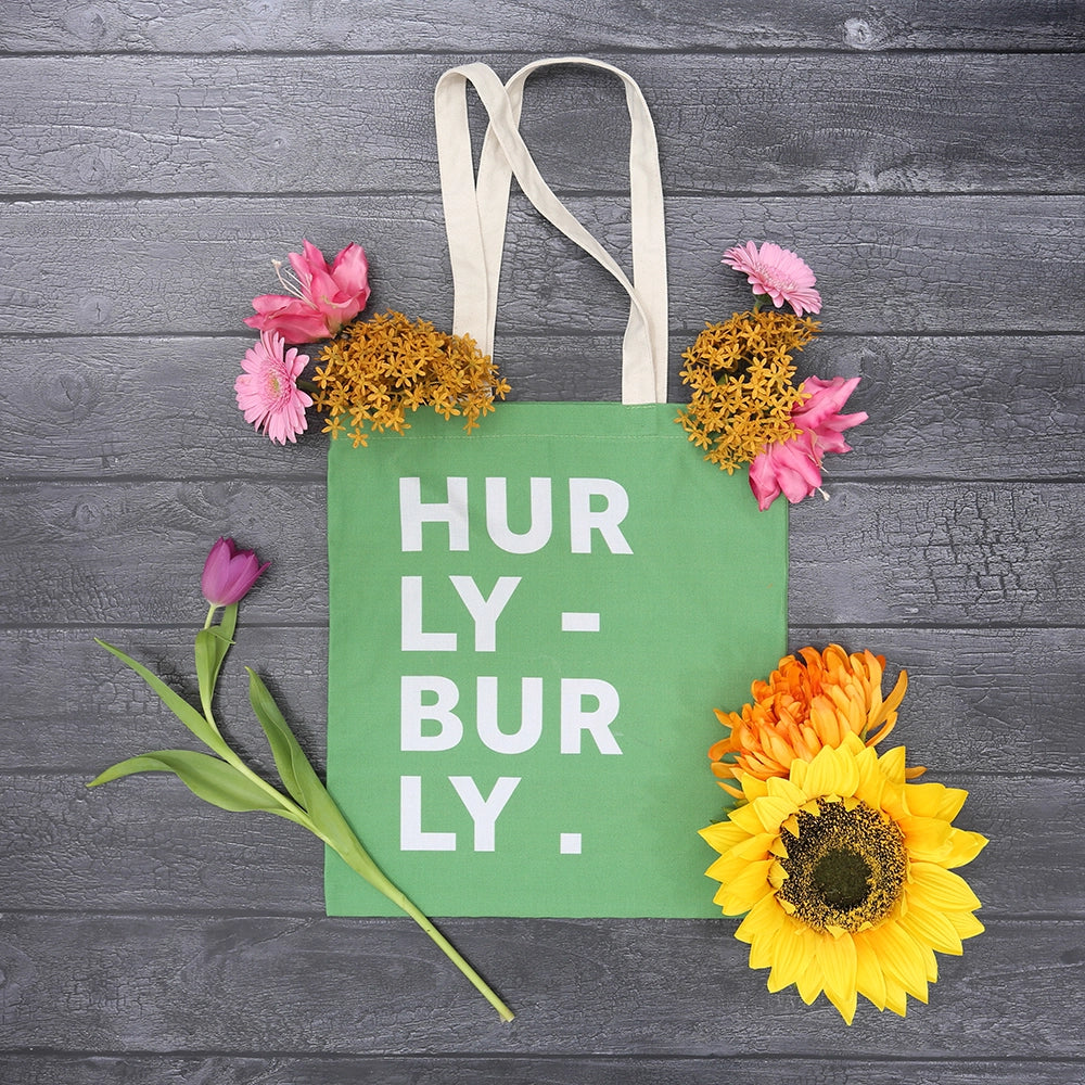 Lime green cotton tote bag with natural handles and white graphic text on front, featuring scattered flowers in background