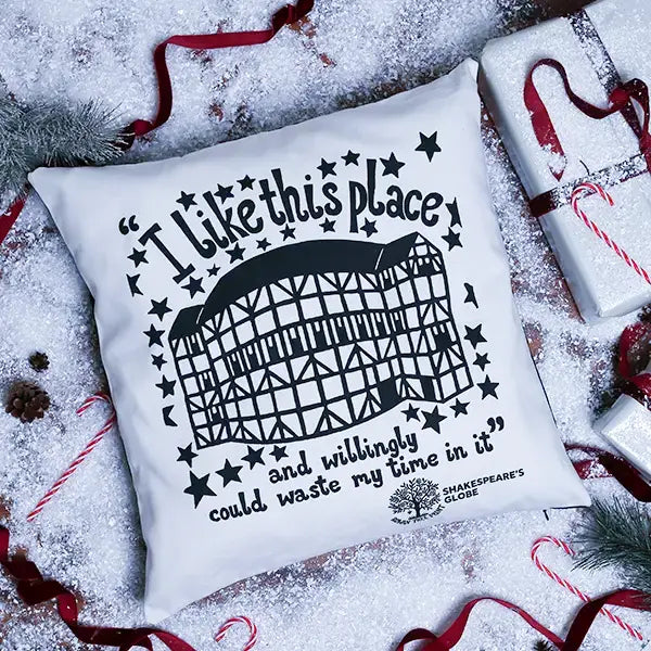 A square white throw pillow with a black graphic of Shakespeare's Globe and the quote 'I like this place and willingly could waste my time in it', styled on a snowy surface with Christmas decorations and wrapped gifts.
