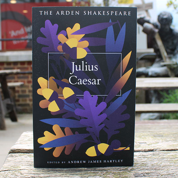 Book titled 'Julius Caesar' by The Arden Shakespeare on a wooden surface with a blurred outdoor background.