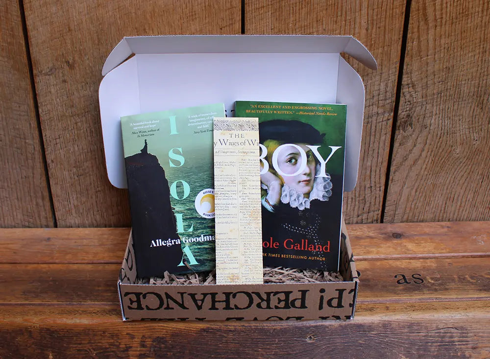 An open cardboard book subscription box filled with shredded paper, featuring Isola by Allegra Goodman, Boy by Nicole Galland, and a Shakespeare-themed bookmark standing upright between the books