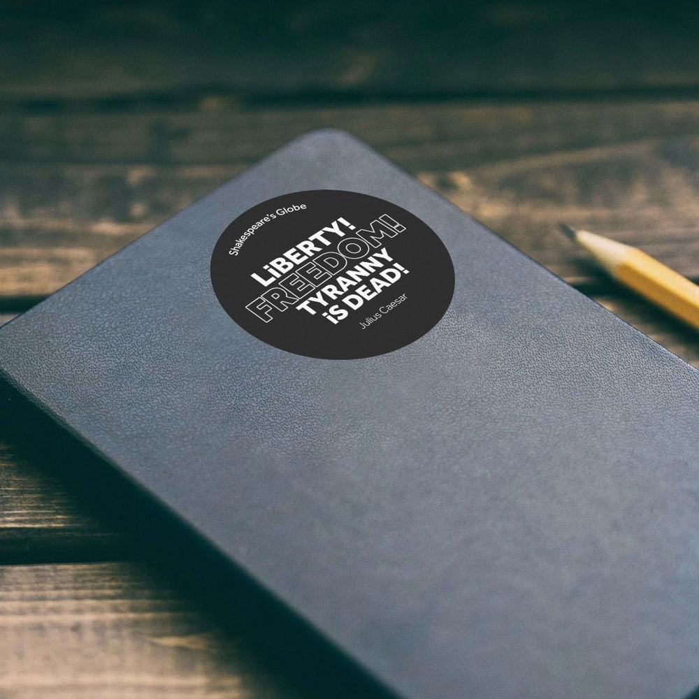 Black circular sticker with white graphic text, stuck to black notebook, sitting on wooden slat surface with sharpened pencil beside it