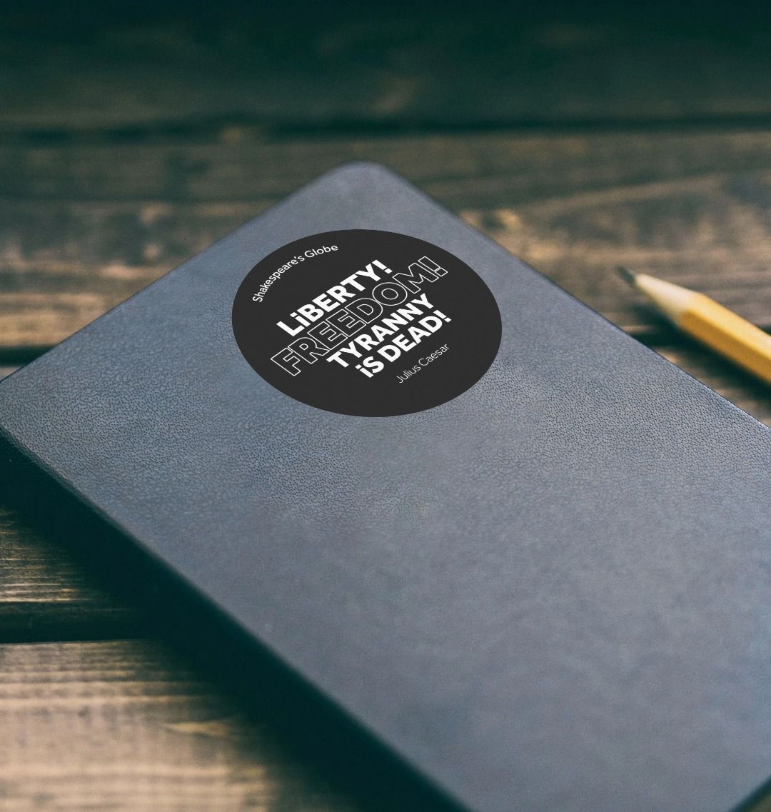 Black circular sticker with white graphic text, stuck to black notebook, sitting on wooden slat surface with sharpened pencil beside it