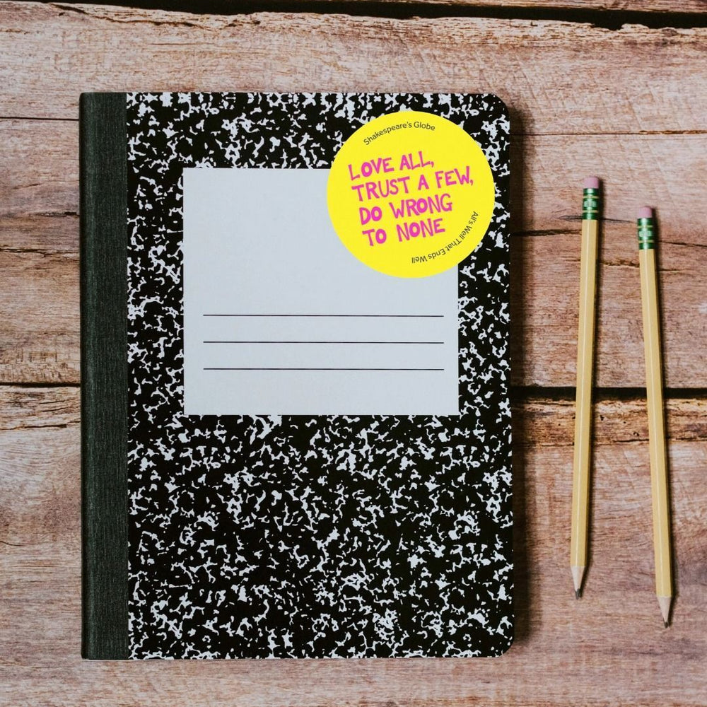 Yellow circular sticker with hot pink graphic text all over print, stuck on retro style notebook which is on wooden slat surface with 2 sharpened pencils sitting beside it