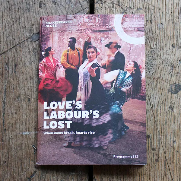 A Shakespeare's Globe programme booklet titled 'Love's Labour's Lost' lies flat on a rustic wooden surface. The cover features a vintage-style photograph of Shakespearean dancers, with the title printed in bold white text.