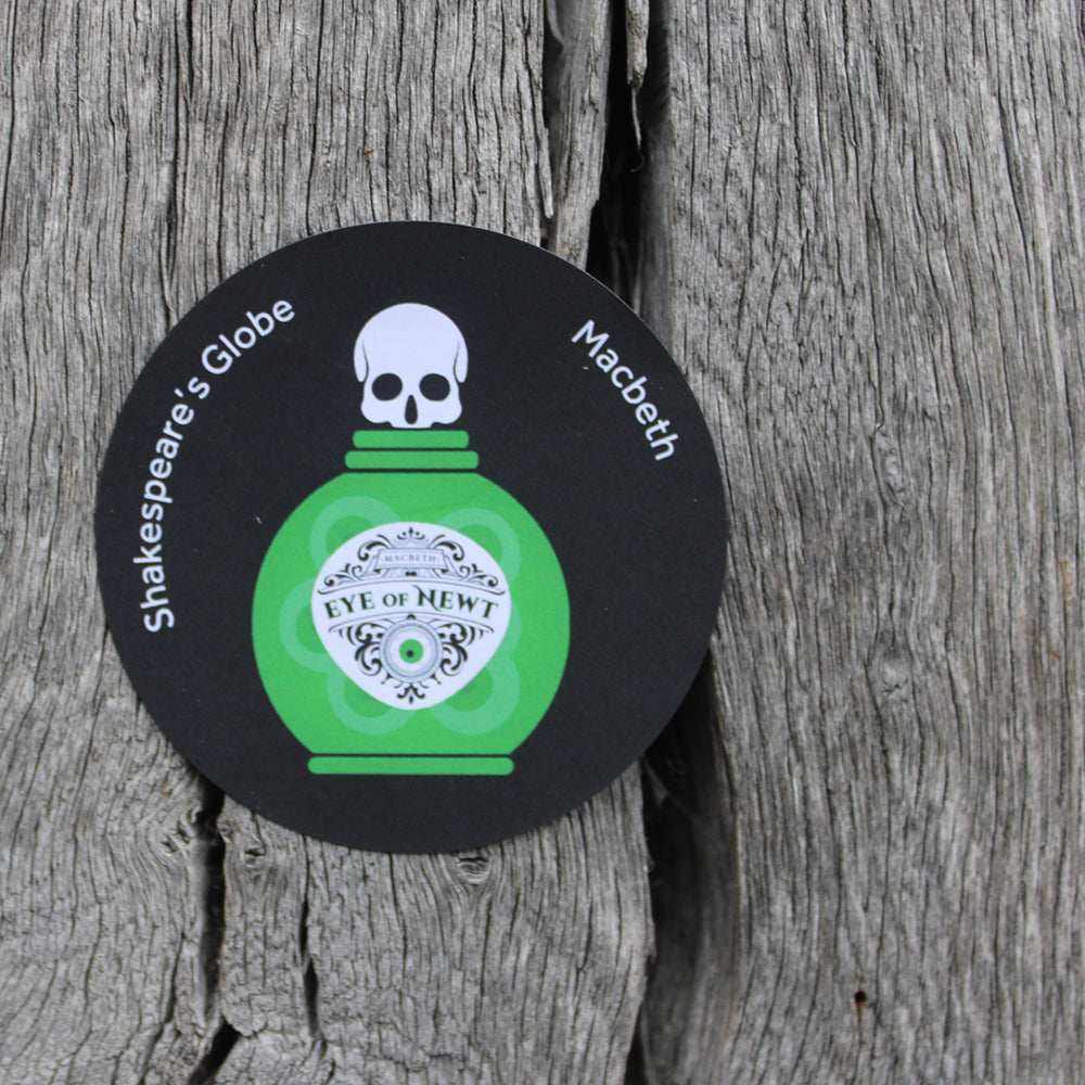 Black circular sticker with green eye of newt bottle graphic on the front, with white text, stuck on wooden surface