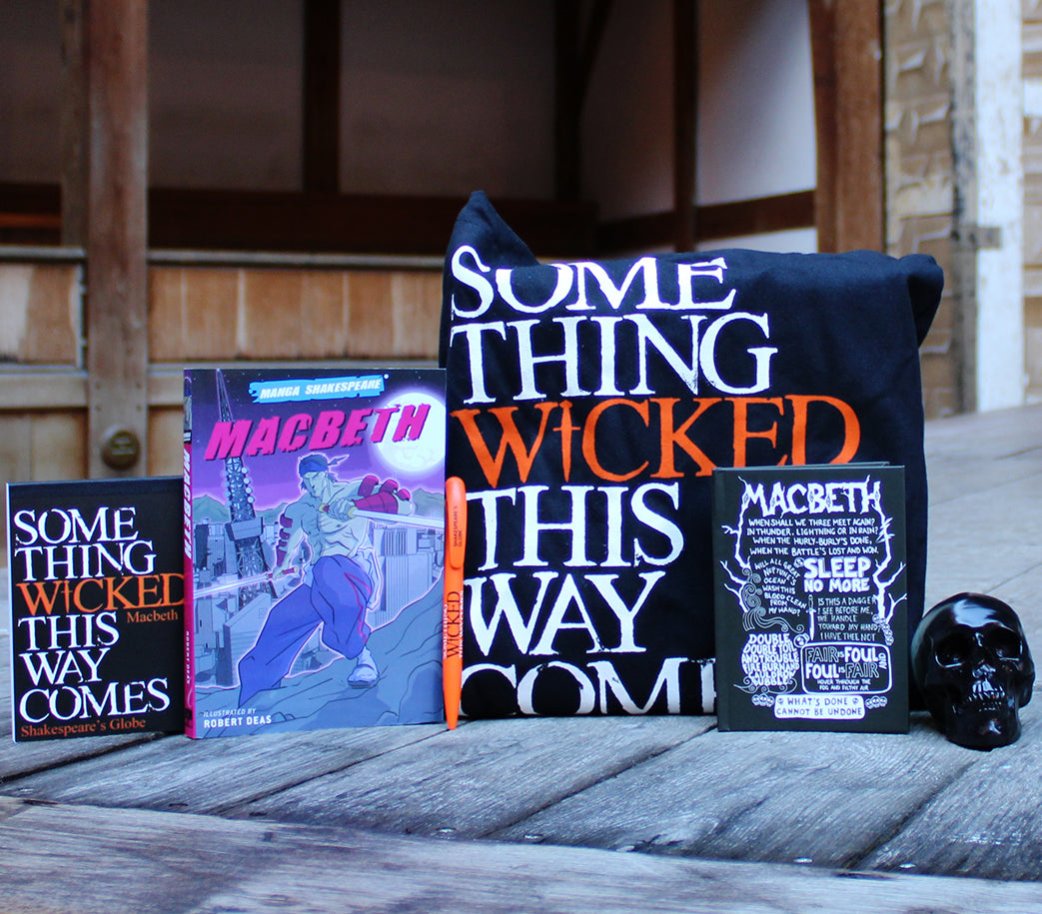 Macbeth themed gifts on wood panel Globe stage, from left to right, paperback notebook. paperback manga, orange pen, black tote bag, hardback notebook, small black prop skull