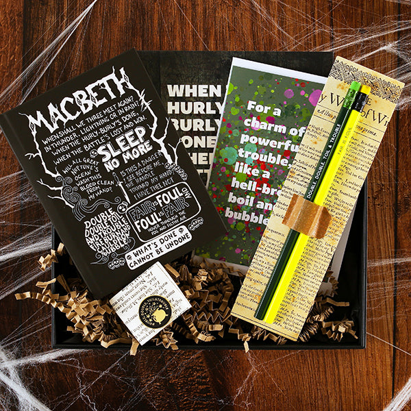 Black box containing pencils on a backing card, a journal with white print, a green greetings card, a grey print and a black and gold pin badge.