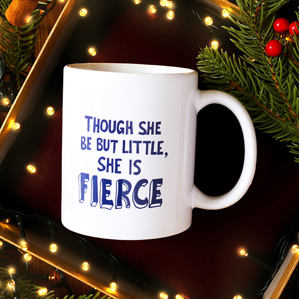 White mug with 'Though she be but little, she is fierce' text on a dark background with Christmas decorations.