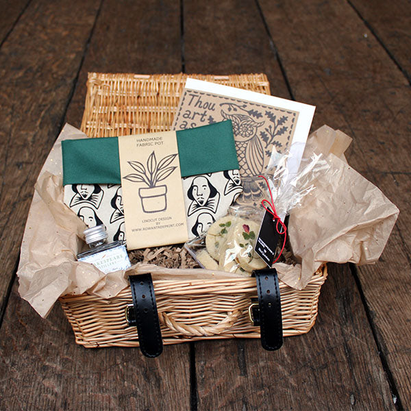 Wicker hamper containing a selection of products, including biscuits, a card, a fabric pot and a bottle