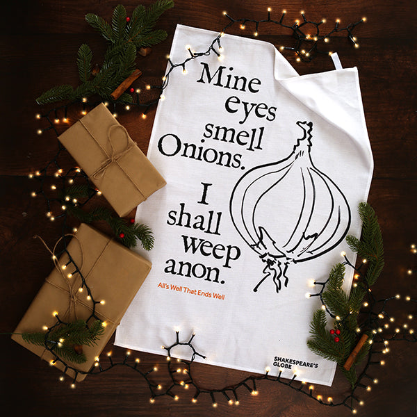 White tea towel with black text and onion illustration on a wooden surface with Christmas decorations.