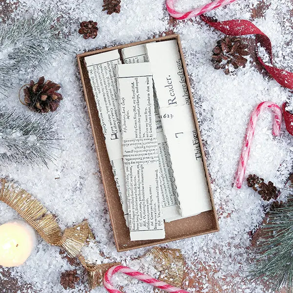 A flat-lay of a brown kraft paper gift box containing vintage-style book pages with printed text, surrounded by festive winter decorations including pinecones, candy canes, a candle, ribbon, and faux snow.