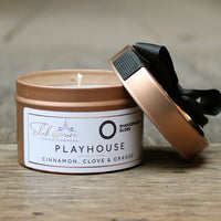 Candle in a rose gold coloured metal container with a black ribbon on a wooden surface, featuring the Shakespeare Globe Theatre label.