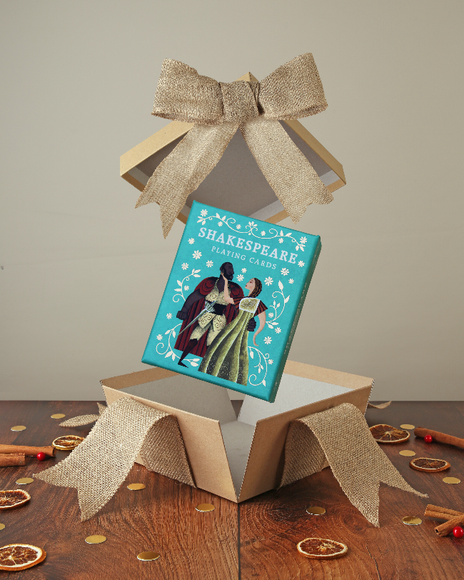 
                      
                        Teal portrait orientated box with male and female figure in Shakespearean dress on cover, popping out of kraft box with bow on wood base with Christmas decor
                      
                    