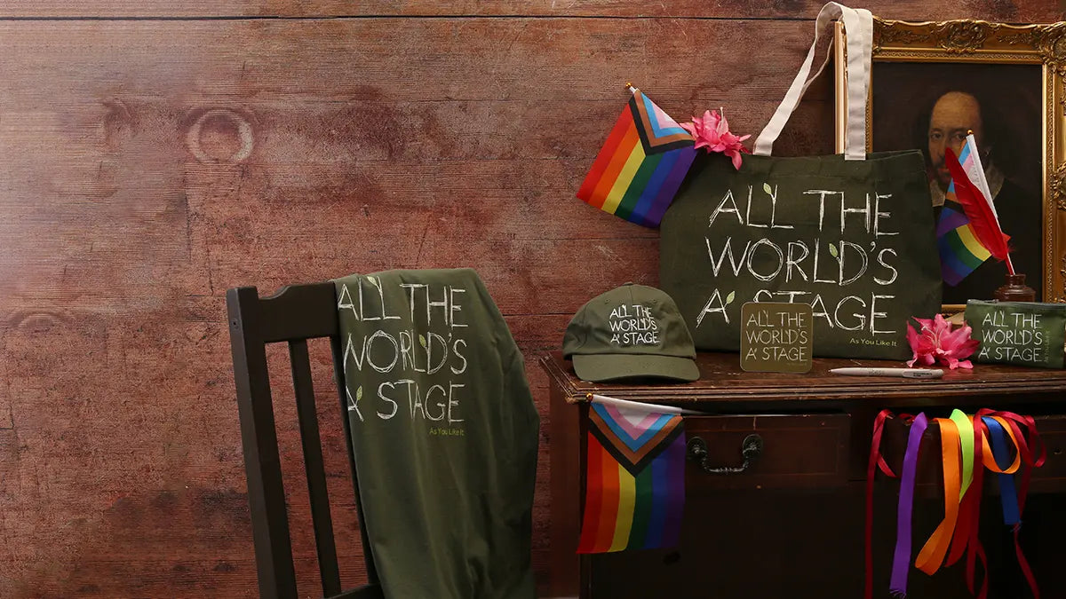Pride-themed merchandise display featuring an olive-green tote bag, T-shirt, cap, mug, and coaster printed with the quote “All the world’s a stage,” arranged on a wooden table with rainbow Pride flags and colourful ribbons.