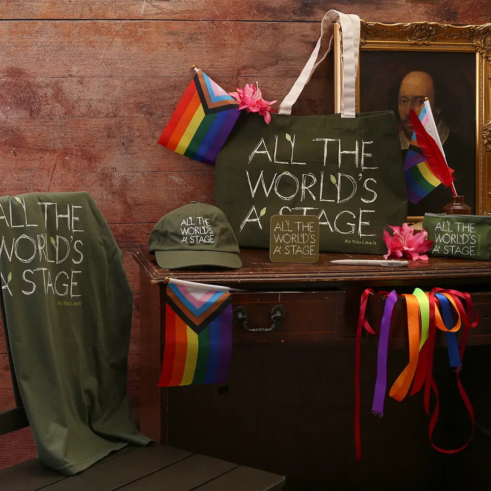 Pride-themed merchandise display featuring an olive-green tote bag, T-shirt, cap, mug, and coaster printed with the quote “All the world’s a stage,” arranged on a wooden table with rainbow Pride flags and colourful ribbons.