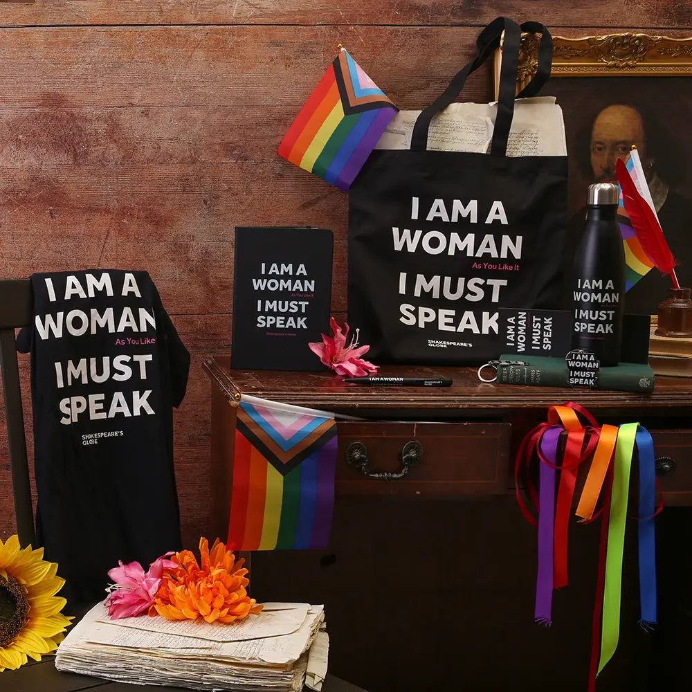Pride-themed merchandise display featuring a black tote bag, T-shirt, notebook, mug, water bottle, and keyring printed with the quote “I am a woman, I must speak,” arranged with rainbow Pride flags and colourful ribbon