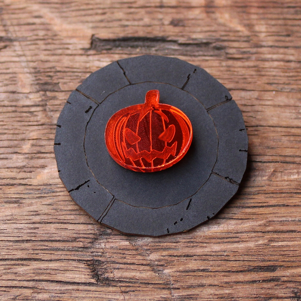 Orange plastic pumpkin pin with etched Jack o'Lantern face in the centre, mounted on matt black card