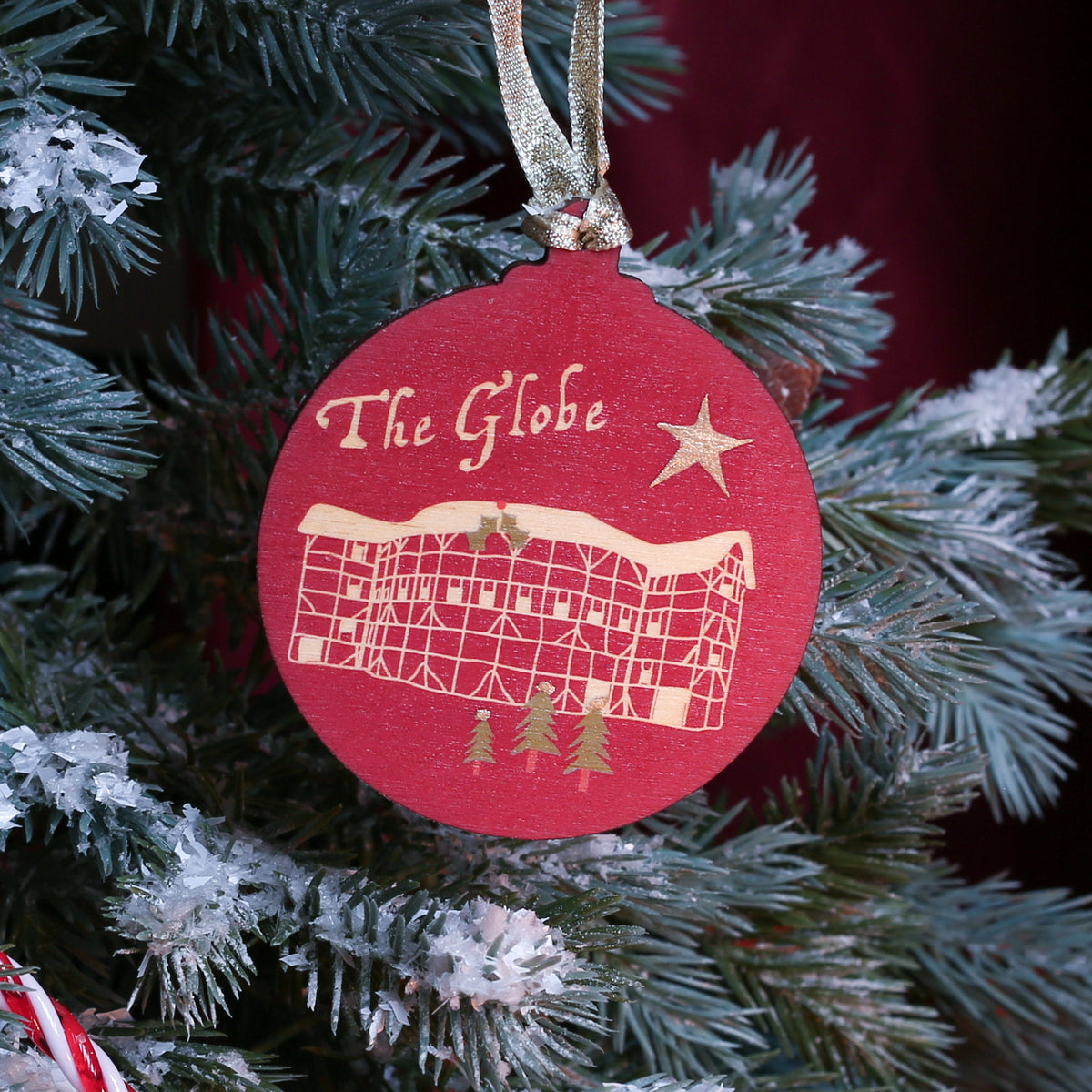 A round red Christmas ornament with gold lettering reading 'The Globe' and a gold illustration of a theatre, hanging on a flocked green Christmas tree with candy canes and a dark red curtain background.
